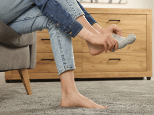 Person seated on a chair/removing a gray sock from a bare foot on a carpet, with a wooden dresser in the background.